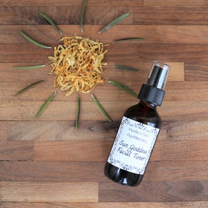 May include: A brown glass bottle with a white label that reads "Herbs & Soil Apothecary Sun Goddess Facial Toner". The bottle is on a wooden surface with dried yellow flower petals and green sprigs arranged in a sun shape.