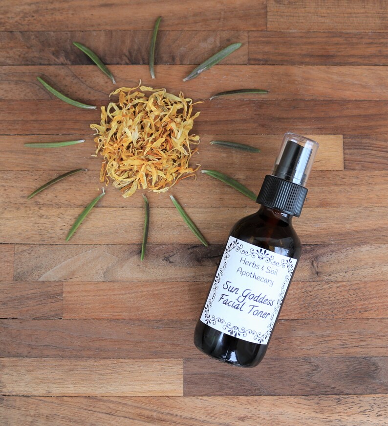 May include: A brown glass bottle with a white label that reads "Herbs & Soil Apothecary Sun Goddess Facial Toner". The bottle is on a wooden surface with dried yellow flower petals and green sprigs arranged in a sun shape.