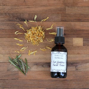 Pode incluir: Uma garrafa de vidro marrom com um bico de pulveriza&ccedil;&atilde;o, etiquetada "Herbs & Soil Apothecary Sun Goddess Facial Toner" sobre uma superf&iacute;cie de madeira com p&eacute;talas de flores amarelas secas e ramos de alecrim verde.