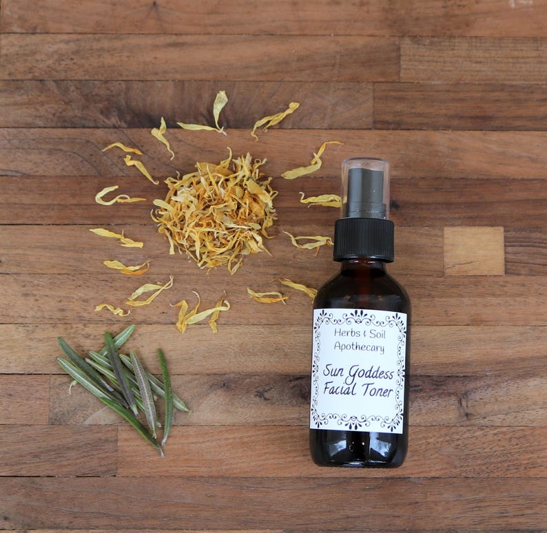 Pode incluir: Uma garrafa de vidro marrom com um bico de pulveriza&ccedil;&atilde;o, etiquetada "Herbs & Soil Apothecary Sun Goddess Facial Toner" sobre uma superf&iacute;cie de madeira com p&eacute;talas de flores amarelas secas e ramos de alecrim verde.