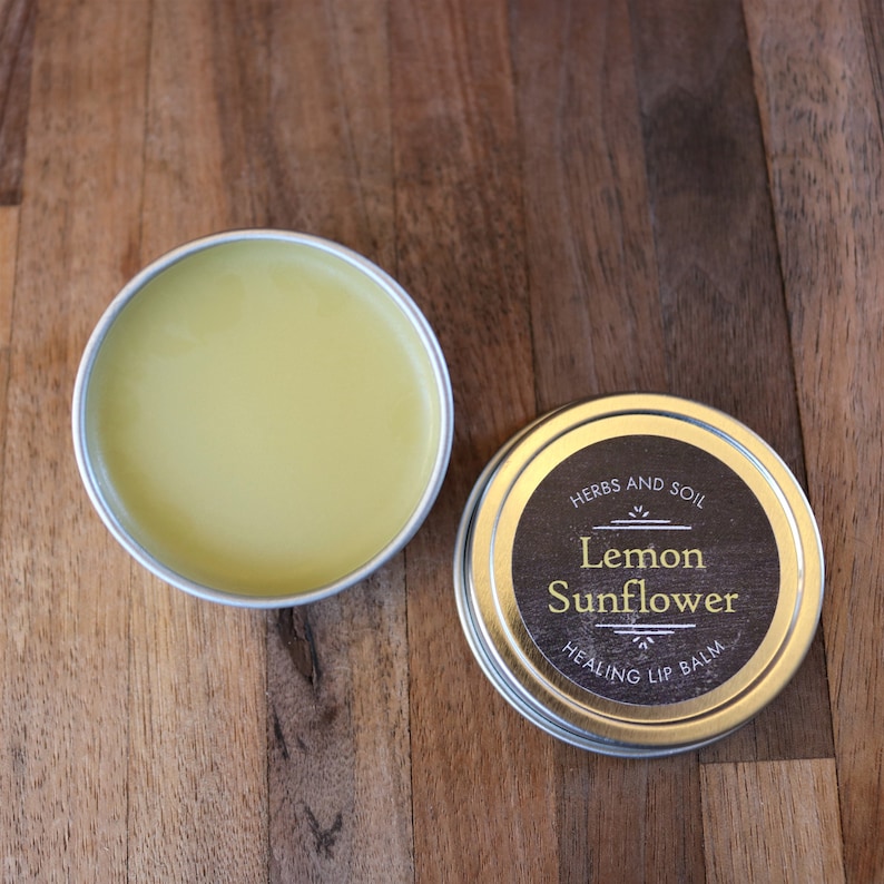 May include: Two silver tins of lip balm, one open and one closed. The open tin reveals yellow balm. The closed tin has a black label with gold text: "Lemon Sunflower Healing Lip Balm" and "Herbs and Soil".