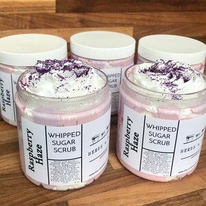 May include: Four clear plastic jars filled with light pink and white whipped sugar scrub, topped with white cream and purple sprinkles. The white labels feature the text Raspberry Haze, Whipped Sugar Scrub, and Heres & S Apothecary with ingredients.