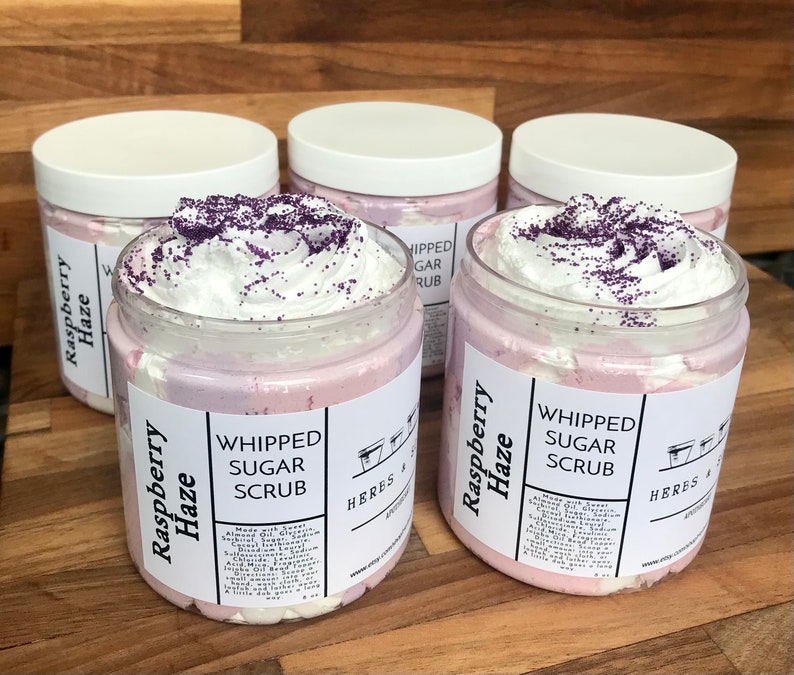 May include: Four clear plastic jars filled with light pink and white whipped sugar scrub, topped with white cream and purple sprinkles. The white labels feature the text Raspberry Haze, Whipped Sugar Scrub, and Heres & S Apothecary with ingredients.
