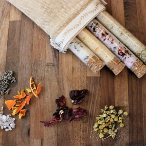 May include: Four glass test tubes with cork stoppers containing various dried botanicals and salts, partially tucked into a beige burlap drawstring pouch with a cream-colored ribbon trim featuring a delicate leaf pattern printed on the fabric.
