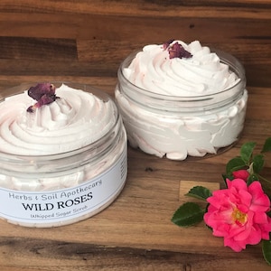 May include: Two jars of whipped sugar scrub with a pink rose petal on top. The jars are labeled "Herbs & Soil Apothecary Wild Roses Whipped Sugar Scrub".