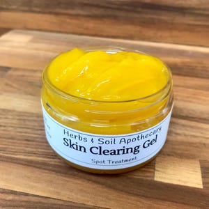 Pode incluir: Um frasco de vidro transparente cheio de um gel transparente para clarear a pele de cor amarela brilhante. O frasco tem um r&oacute;tulo branco que diz "Herbs & Soil Apothecary Skin Clearing Gel Spot Treatment".