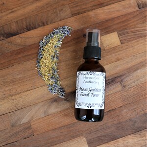 May include: A brown glass bottle with a black spray top and a white label that reads "Herbs & Soil Apothecary Moon Goddess Facial Toner". The bottle is sitting on a wooden surface with dried lavender and yellow flowers.