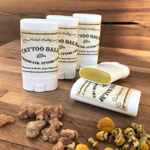 May include: Four white plastic twist-up tubes of tattoo balm with cream-colored labels. The labels feature the text Herbal Healing, TATTOO BALM, FRESH INK AFTERCARE, and Herbs & Soil Apothecary. One tube is lying on its side with the balm exposed.