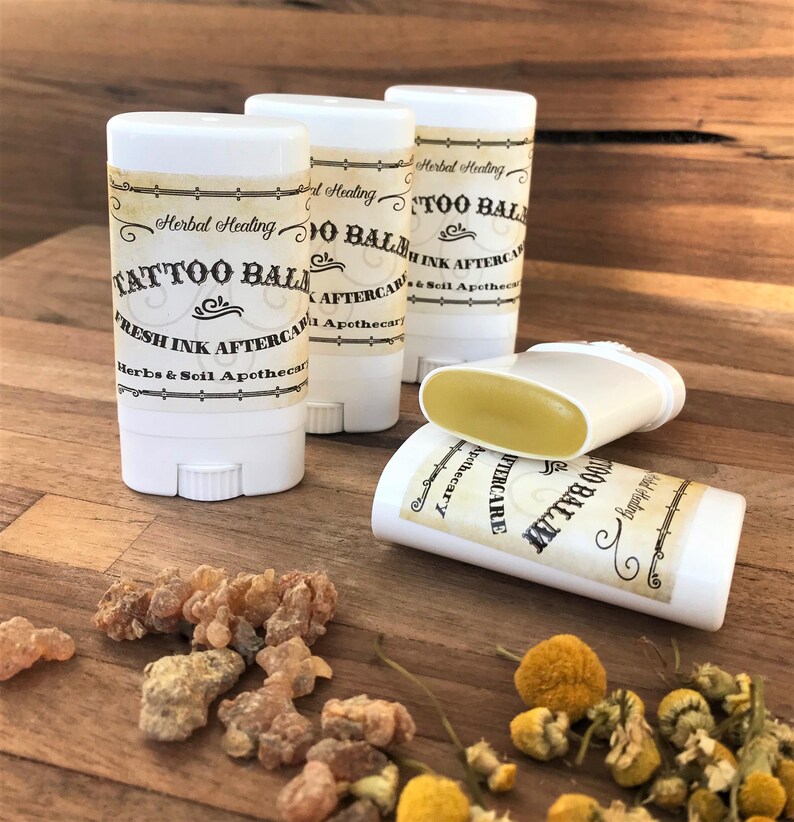 May include: Four white plastic twist-up tubes of tattoo balm with cream-colored labels. The labels feature the text Herbal Healing, TATTOO BALM, FRESH INK AFTERCARE, and Herbs & Soil Apothecary. One tube is lying on its side with the balm exposed.