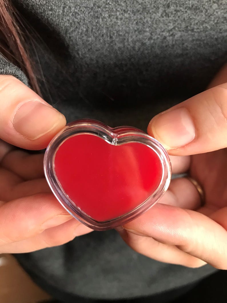 Pode incluir: Um recipiente em forma de cora&ccedil;&atilde;o com b&aacute;lsamo labial vermelho dentro. O recipiente &eacute; de pl&aacute;stico transparente e o b&aacute;lsamo labial &eacute; de cor vermelha brilhante.