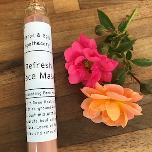 Pode incluir: Um pequeno frasco de vidro com tampa de corti&ccedil;a cheio de p&oacute; de m&aacute;scara facial rosa. O frasco tem um r&oacute;tulo branco com o texto "Herbs & Soil Apothecary" e "Refresh Face Mask" impresso nele. O frasco est&aacute; sentado em uma superf&iacute;cie de madeira com duas rosas cor-de-rosa.