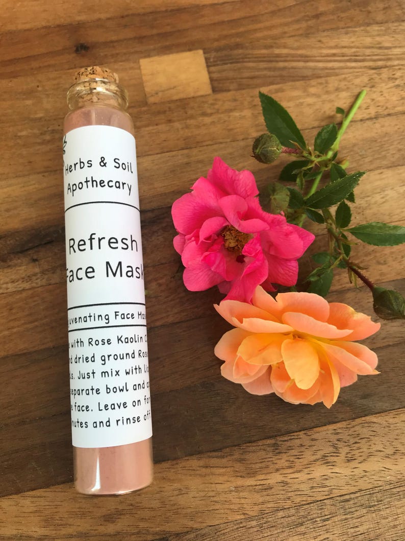 Pode incluir: Um pequeno frasco de vidro com tampa de corti&ccedil;a cheio de p&oacute; de m&aacute;scara facial rosa. O frasco tem um r&oacute;tulo branco com o texto "Herbs & Soil Apothecary" e "Refresh Face Mask" impresso nele. O frasco est&aacute; sentado em uma superf&iacute;cie de madeira com duas rosas cor-de-rosa.
