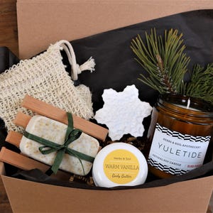 May include: A gift box filled with bath and body products, including a natural soy candle labelled "Yuletide", a bar of soap, a wooden soap dish, a small jar of body butter labelled "Warm Vanilla", and a white snowflake ornament.
