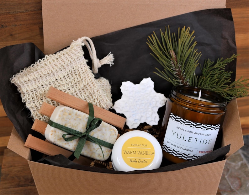 May include: A gift box filled with bath and body products, including a natural soy candle labelled "Yuletide", a bar of soap, a wooden soap dish, a small jar of body butter labelled "Warm Vanilla", and a white snowflake ornament.