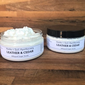 May include: Two white jars of whipped sugar scrub with black lids. The jars are labeled "Herbs & Soil Apothecary Leather & Cedar Whipped Sugar Scrub".