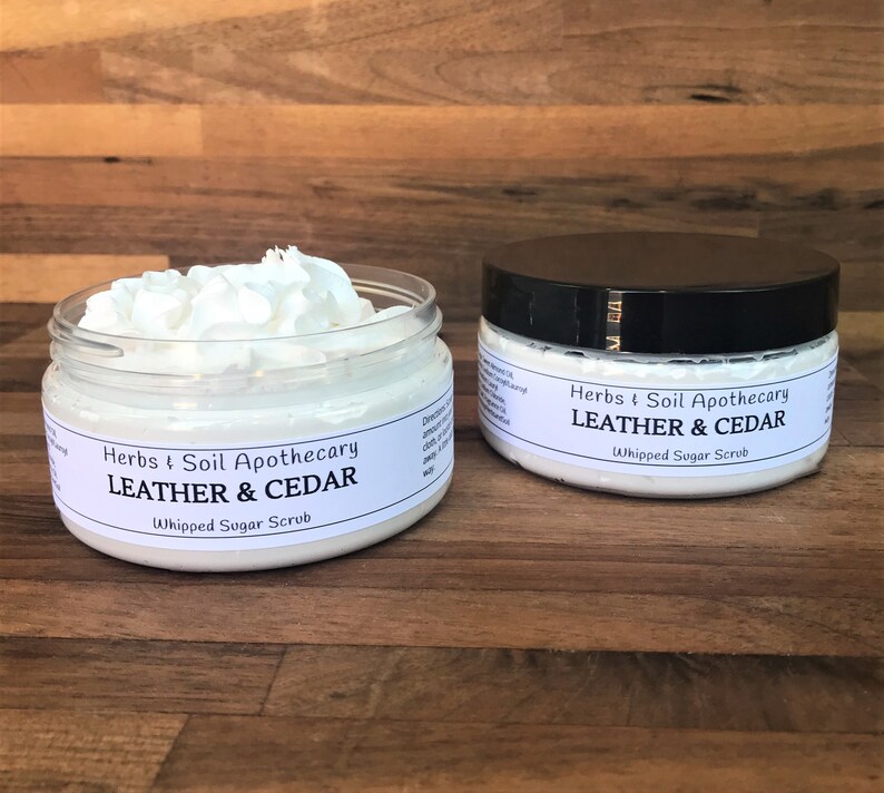 May include: Two white jars of whipped sugar scrub with black lids. The jars are labeled "Herbs & Soil Apothecary Leather & Cedar Whipped Sugar Scrub".