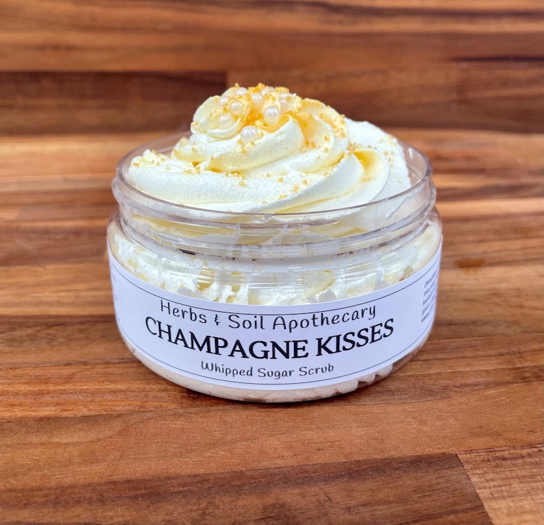 May include: A clear jar filled with a whipped sugar scrub. The scrub is topped with swirls of white cream and decorated with small, gold-colored sprinkles and white pearl-like beads. The label reads "Champagne Kisses" and "Whipped Sugar Scrub."