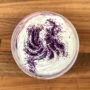 May include: A clear plastic container filled with a thick, white, whipped substance that has a swirled texture. The top surface is decorated with a generous dusting of small, round, deep purple sprinkles arranged in a swirling pattern across the cream.