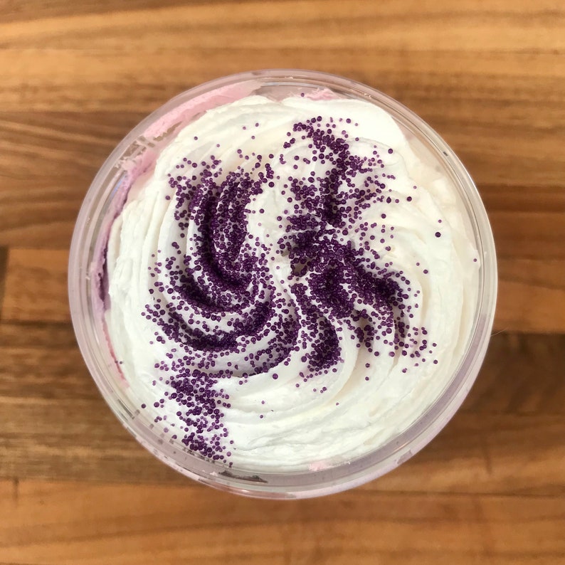 May include: A clear plastic container filled with a thick, white, whipped substance that has a swirled texture. The top surface is decorated with a generous dusting of small, round, deep purple sprinkles arranged in a swirling pattern across the cream.