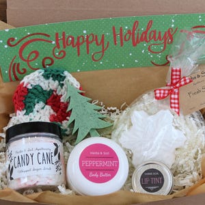 May include: A gift box containing a jar of Candy Cane whipped sugar scrub, a jar of Peppermint body butter, a small tin of lip tint, a white snowflake-shaped soap, a green tree-shaped soap, and a crocheted round pad with a Happy Holidays sign.