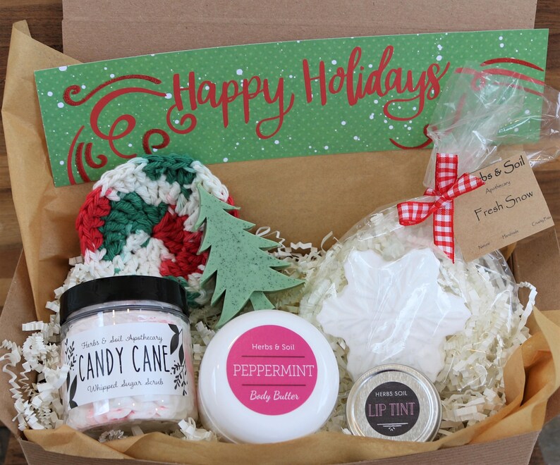May include: A gift box containing a jar of Candy Cane whipped sugar scrub, a jar of Peppermint body butter, a small tin of lip tint, a white snowflake-shaped soap, a green tree-shaped soap, and a crocheted round pad with a Happy Holidays sign.