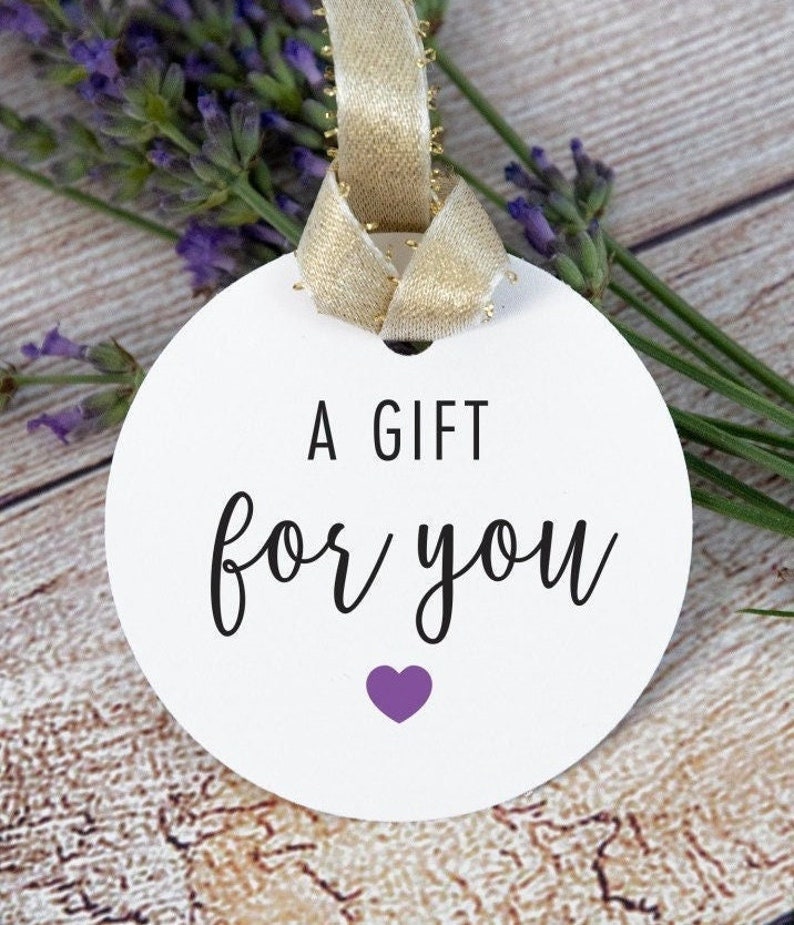 May include: A white round gift tag with a gold ribbon attached. The tag has black text that says "A Gift for You" and a small purple heart.