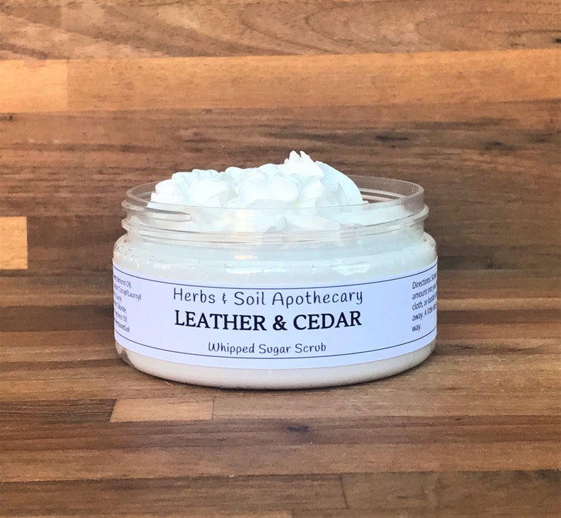 May include: A white jar of whipped sugar scrub with a white label that reads "Herbs & Soil Apothecary Leather & Cedar Whipped Sugar Scrub".