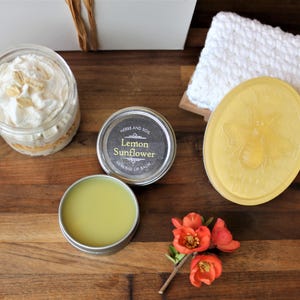 May include: A gift basket containing a jar of whipped body butter, a tin of lemon sunflower healing lip balm, a bar of yellow soap with a bee design, and a white crocheted washcloth.