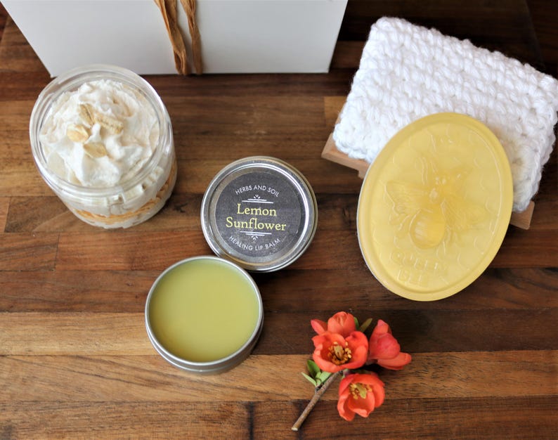 May include: A gift basket containing a jar of whipped body butter, a tin of lemon sunflower healing lip balm, a bar of yellow soap with a bee design, and a white crocheted washcloth.