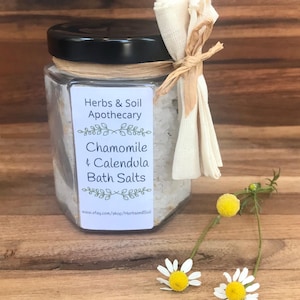 Pode incluir: Um frasco de vidro cheio de sais de banho brancos, amarrado com uma fita marrom e um saco de tecido branco. O r&oacute;tulo diz "Herbs & Soil Apothecary, Chamomile & Calendula Bath Salts, www.etsy.com/shop/HerbsandSoil". Tr&ecirc;s margaridas brancas com centros amarelos est&atilde;o em primeiro plano.