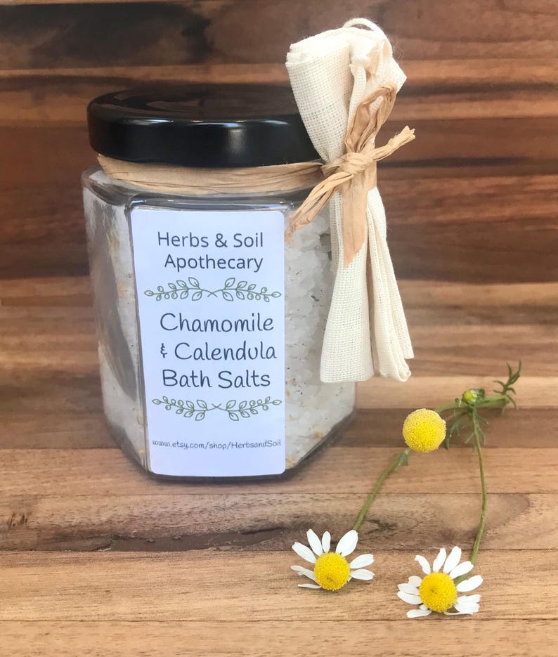 Pode incluir: Um frasco de vidro cheio de sais de banho brancos, amarrado com uma fita marrom e um saco de tecido branco. O r&oacute;tulo diz "Herbs & Soil Apothecary, Chamomile & Calendula Bath Salts, www.etsy.com/shop/HerbsandSoil". Tr&ecirc;s margaridas brancas com centros amarelos est&atilde;o em primeiro plano.