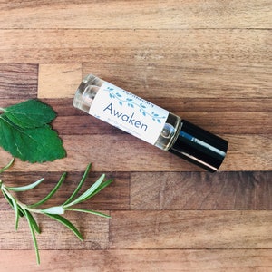 May include: A clear glass roll-on bottle with a black cap. The label is white with green vines and the text "Apothecary Awaken Roll On Aromatherapy".