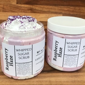 May include: Two jars of whipped sugar scrub in the scent Raspberry Haze. The jars are white with a pink and white swirl design. The label reads "Whipped Sugar Scrub" and "Raspberry Haze".