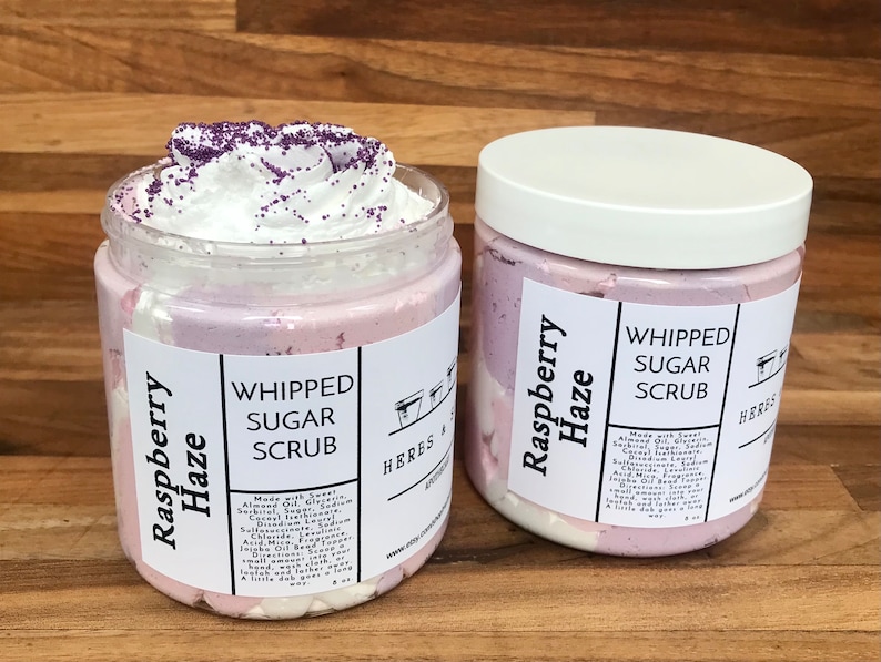 May include: Two clear plastic jars of Raspberry Haze whipped sugar scrub. One jar is open, revealing white whipped scrub topped with purple sprinkles. The other jar is closed with a white lid. Labels feature black text and ingredient lists.