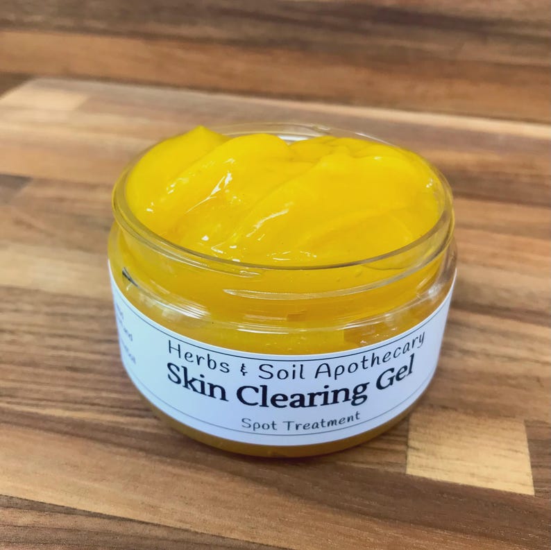 May include: A clear, round plastic jar filled with a vibrant yellow gel. The white label on the front features black text that reads Herbs & Soil Apothecary Skin Clearing Gel Spot Treatment, with a simple design and clear, legible typography.