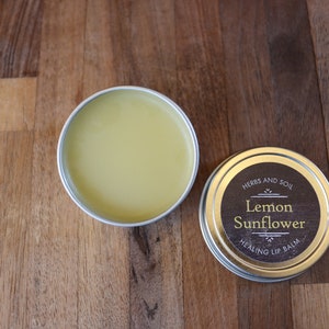May include: Two small, round tins of lip balm on a wooden surface. One tin is open, revealing a yellow balm. The other is closed, with a label reading "Lemon Sunflower Healing Lip Balm".
