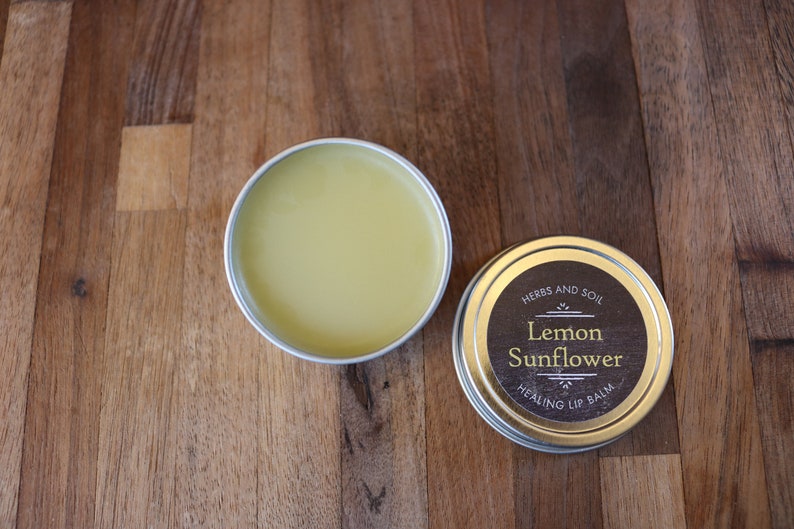 May include: Two small, round tins of lip balm on a wooden surface. One tin is open, revealing a yellow balm. The other is closed, with a label reading "Lemon Sunflower Healing Lip Balm".