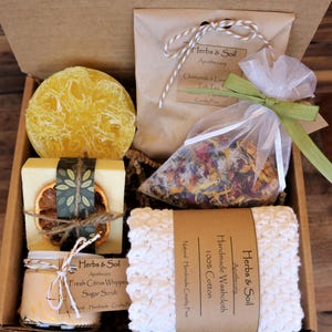 May include: A gift box filled with bath and body products. The box contains a loofah, a bar of soap, a jar of sugar scrub, a bag of bath salts, and a crocheted washcloth. The products are all labelled with the name "Herbs & Soil Apothecary".
