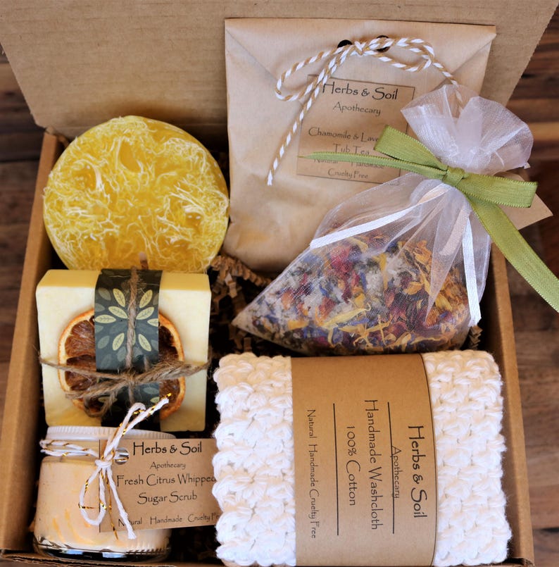 May include: A gift box filled with bath and body products. The box contains a loofah, a bar of soap, a jar of sugar scrub, a bag of bath salts, and a crocheted washcloth. The products are all labelled with the name "Herbs & Soil Apothecary".