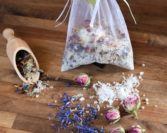 May include: A white organza bag filled with bath salts and dried flowers, including rose petals, lavender, and calendula. The bag is tied with a green ribbon and sits on a wooden surface with loose bath salts and dried flowers scattered around it.