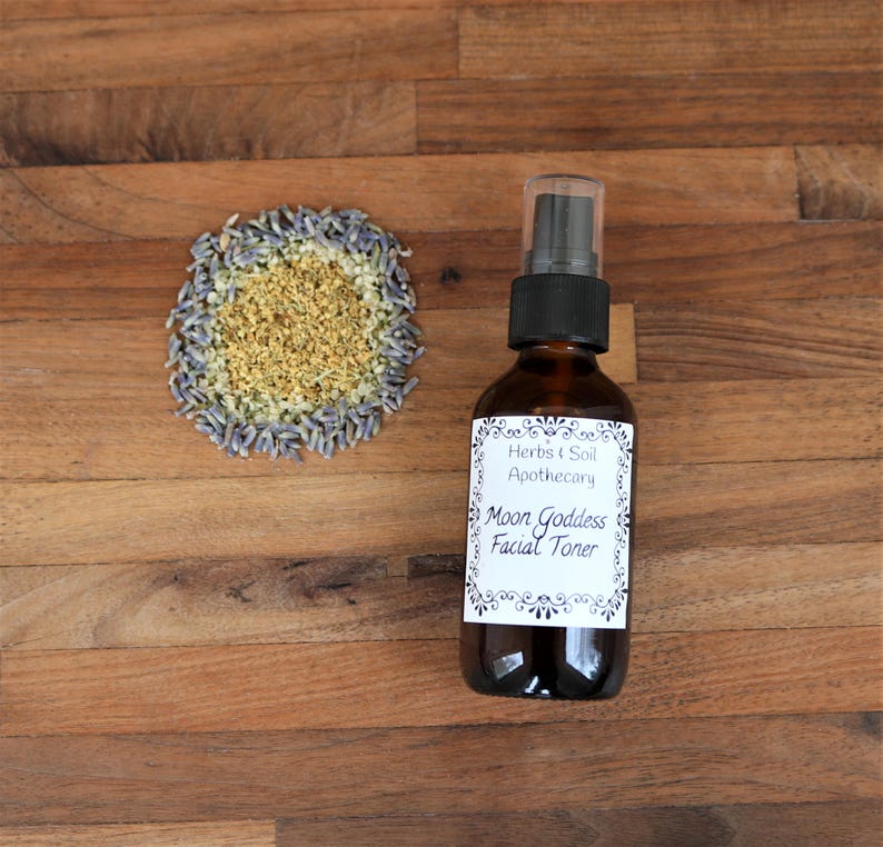 May include: An amber glass spray bottle with a black pump top and a white label reading Herbs & Soil Apothecary Moon Goddess Facial Toner, accompanied by a circular arrangement of dried lavender buds and small yellow botanical flower petals.