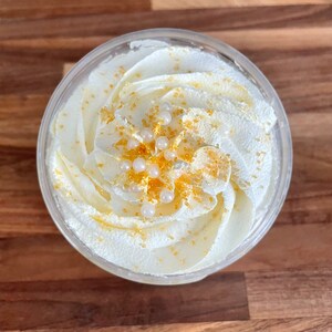 May include: A clear plastic container filled with white whipped cream, swirled and topped with gold sprinkles and white pearl-like decorations. The dessert sits on a wooden surface.