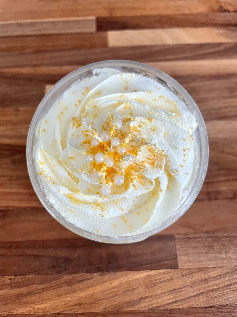 May include: A clear plastic container filled with white whipped cream, swirled and topped with gold sprinkles and white pearl-like decorations. The dessert sits on a wooden surface.