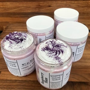 May include: Four clear plastic jars filled with light pink whipped sugar scrub, topped with white cream and purple sprinkles. Each jar features a white label with the text Raspberry Haze, Whipped Sugar Scrub, and Herbs & Soil Apothecary.
