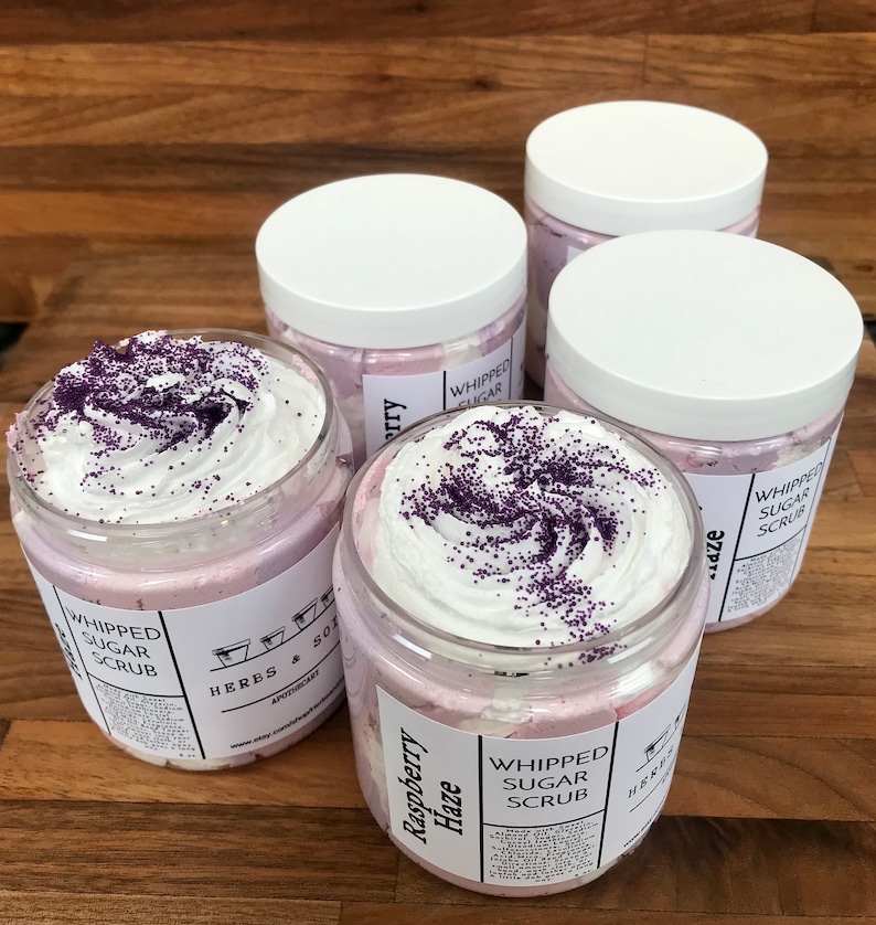 May include: Four clear plastic jars filled with light pink whipped sugar scrub, topped with white cream and purple sprinkles. Each jar features a white label with the text Raspberry Haze, Whipped Sugar Scrub, and Herbs & Soil Apothecary.