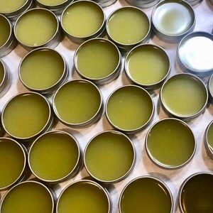 May include: A high-angle view of many small, round, silver tins containing a light green, semi-solid product. The tins are packed closely together, forming a grid. The contents suggest a balm or salve.