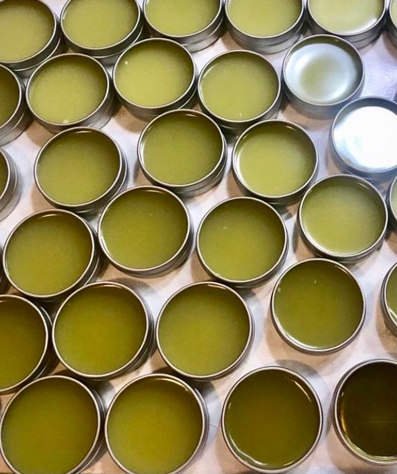 May include: A high-angle view of many small, round, silver tins containing a light green, semi-solid product. The tins are packed closely together, forming a grid. The contents suggest a balm or salve.