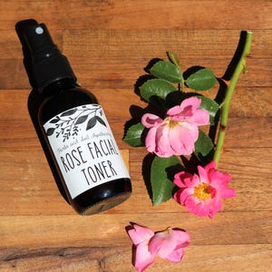 May include: A dark glass bottle with a black spray nozzle and a white label that reads "Herbs and Soil Apothecary Rose Facial Toner". The bottle is sitting on a wooden surface with pink rose petals and a rose stem with green leaves.