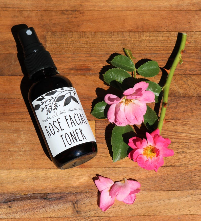 May include: A dark glass bottle with a black spray nozzle and a white label that reads "Herbs and Soil Apothecary Rose Facial Toner". The bottle is sitting on a wooden surface with pink rose petals and a rose stem with green leaves.