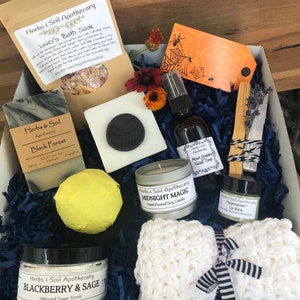 May include: A Halloween-themed gift box filled with bath and body products from Herbs & Soil Apothecary. The box includes a black forest soap, a blackberry and sage whipped sugar scrub, a midnight magic soy candle, a moon goddess facial toner, a nightshade lip balm, a bath bomb, a crocheted washcloth, and a wooden stick of incense.
